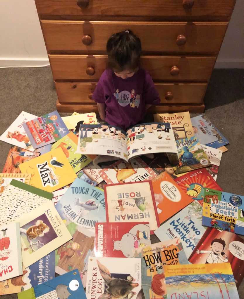 A little girl sits on the floor, surrounded by a pile a books