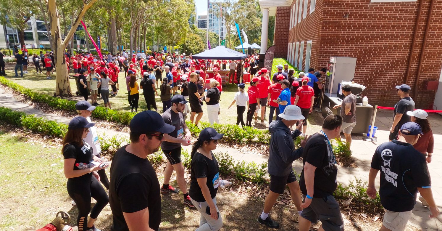 Walkers from Macquarie Park Business Community Partnership returning to the finish line in the shade, on the J&J Lawns for the end of Walkathon BBQ, served on the lawns.